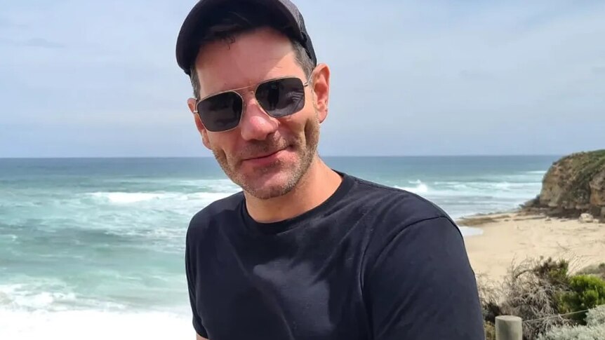 A man in a cap and sunglasses smiles and poses at the beach