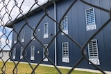 A blue steel clad building, with barred windows, is pictured behind a chain wire fence.