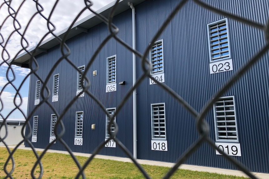 A blue steel clad building, with barred windows, is pictured behind a chain wire fence.