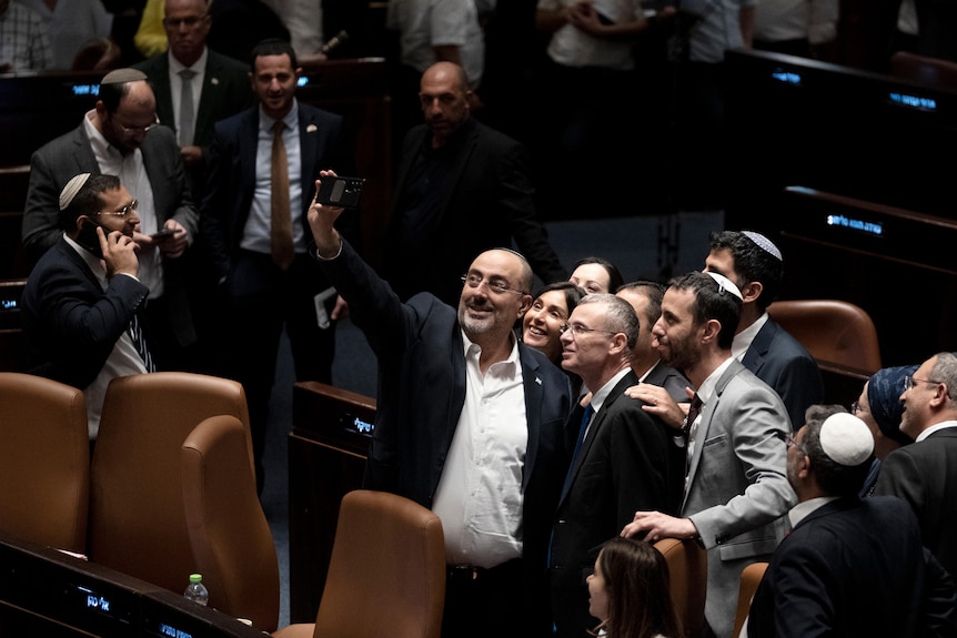 A group of people take a selfie together in a parliamentary chamber.