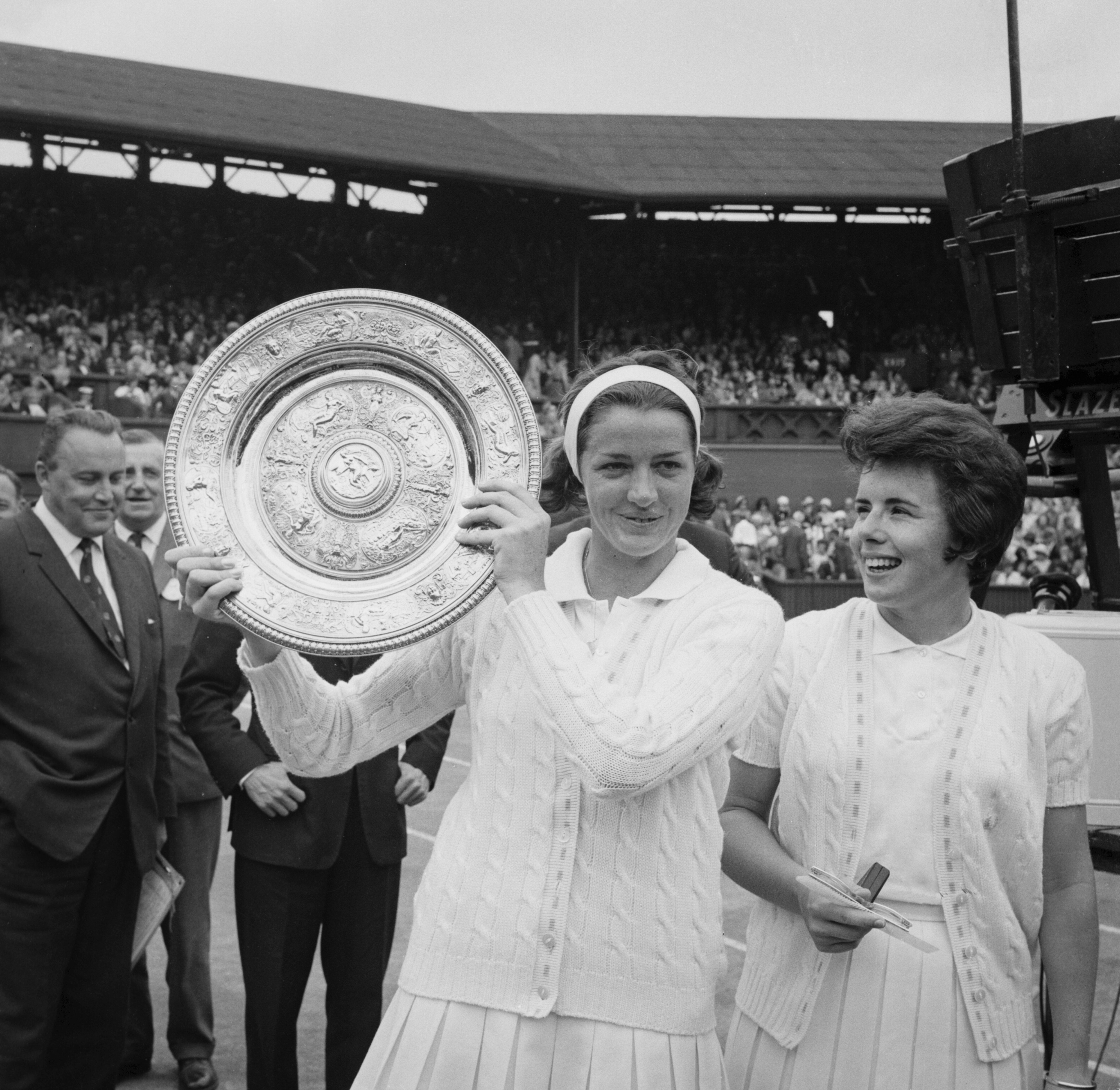 A black and white photo of one tennis player holding a trophy aloft, another next to her.