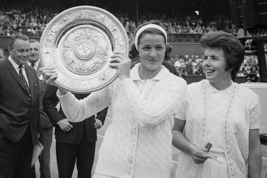 A black and white photo of one tennis player holding a trophy aloft, another next to her.