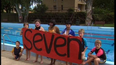Children stand in front of swimming pool holding sign that reads seven