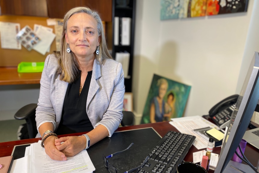 A fair-haired woman sits at a desk with her hands clasped.