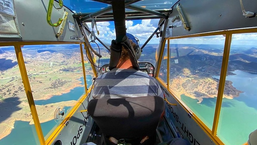 A photo taken inside a plane of a man in a striped shirt operating a plane with large panel windows flying over hills and coast.