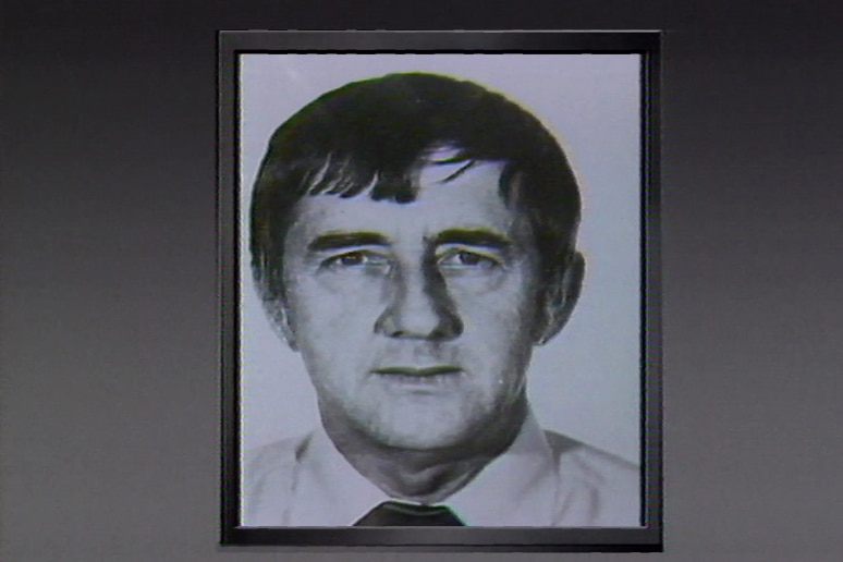 A black and white mugshot of a man with grey hair looking at the camera.