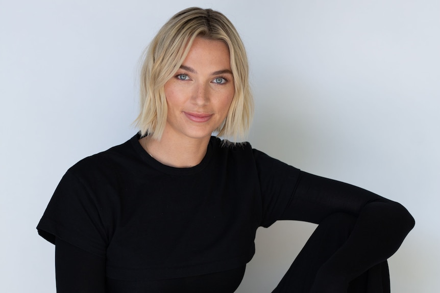 A blonde woman with short hair sits in an all black outfit against a white wall on a stool and half smiles at the camera. 