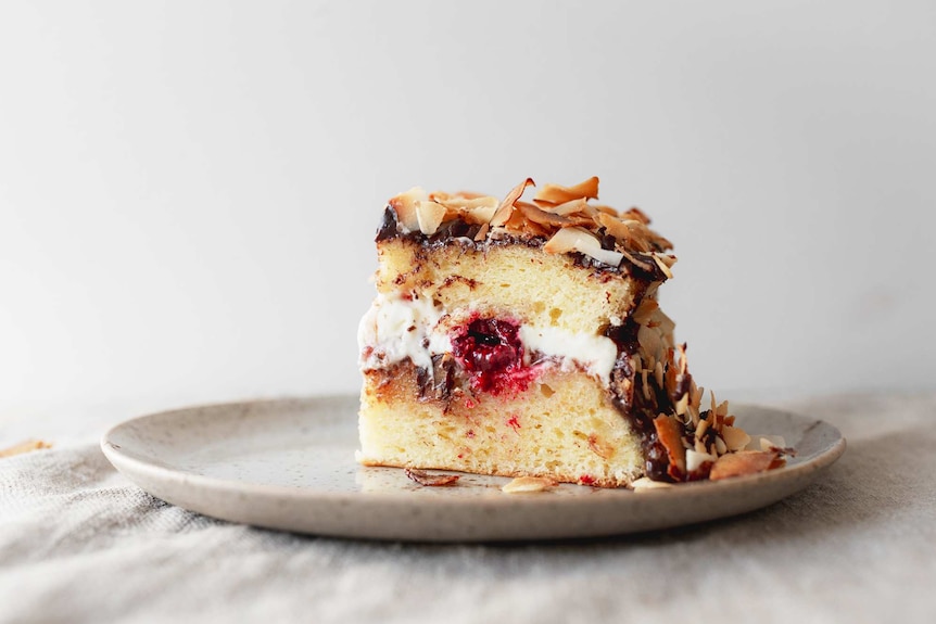 Slice of lamington cake with toasted coconut, cream, jam, fresh raspberries and chocolate ganache on a plate.