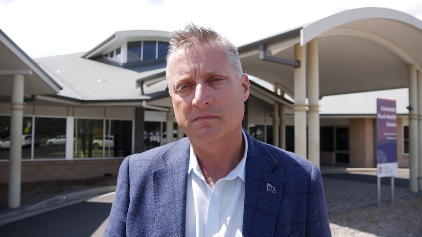 man in a suit standing in front of a health centre. Sombre face.