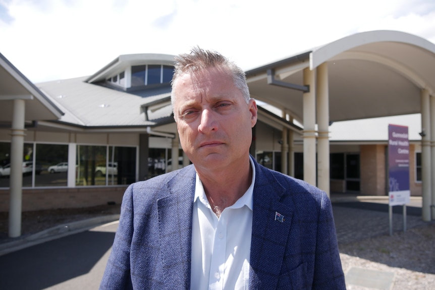 man in a suit standing in front of a health centre. Sombre face.