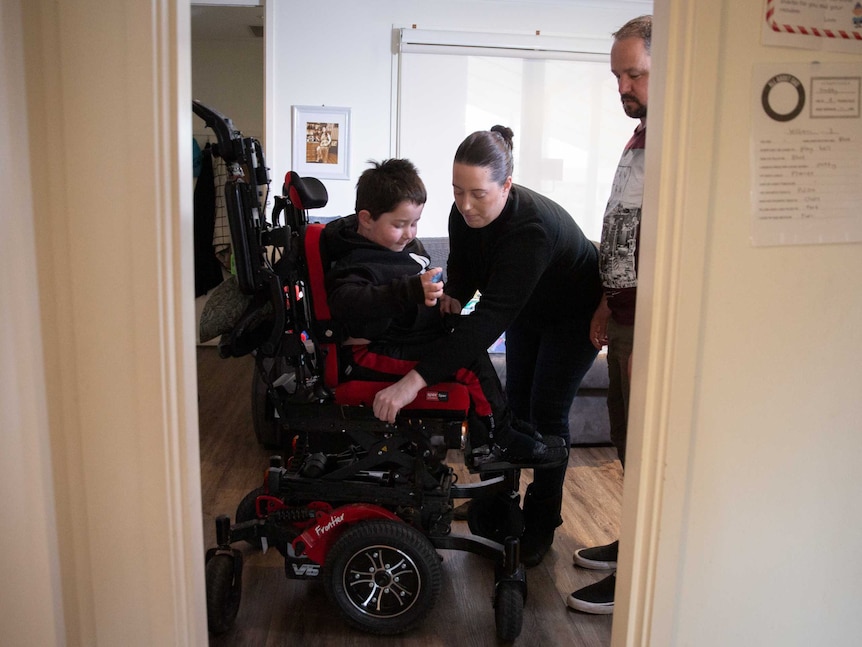 A young boy is helped into his specialist wheelchair by his mum.