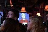 American voters watch the United States Presidential debate on a television screen in a dimly lit bar.