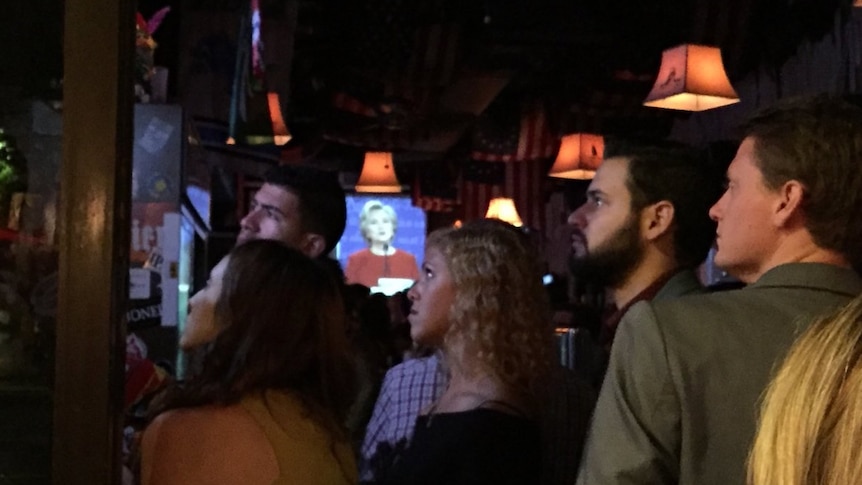 American voters watch the United States Presidential debate on a television screen in a dimly lit bar.