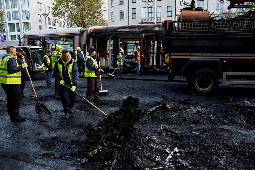 Workers wearing yellow vests scoop up burned asphalt with shovels.