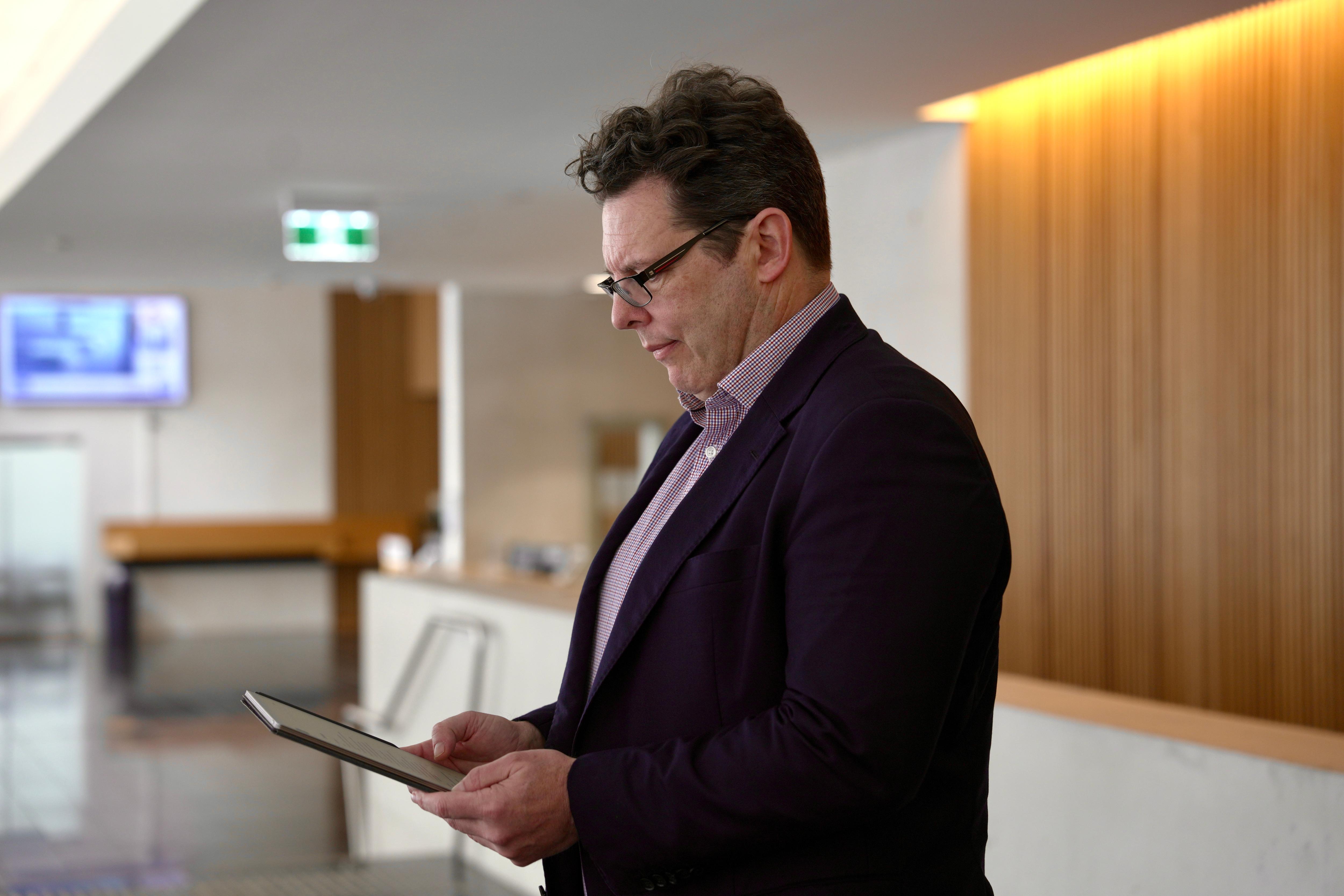 Alex Robson, wearing a dark suit jacket and a collared shirt, looks down at a device in his hands. Behind him is a wooden wall.