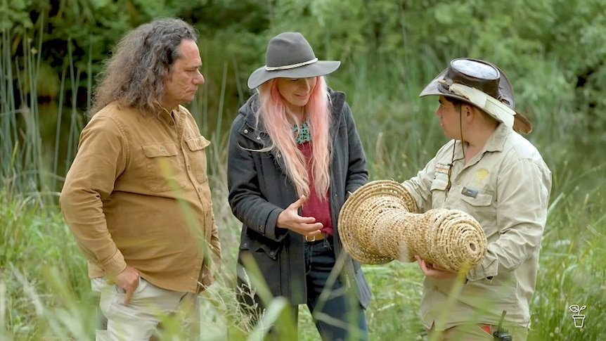 Man showing a woven eel trap to man and woman in bush.
