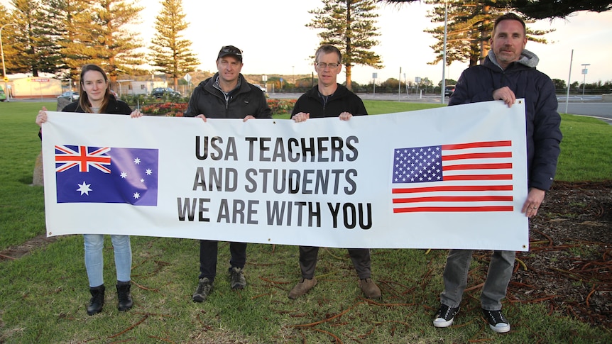 Four people hold up a white banner with the Australian and US flags, reading 'USA teachers and students we are with you'