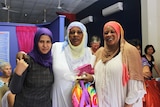 Three women in Arabic headdress smile at a camera in a busy foyer.