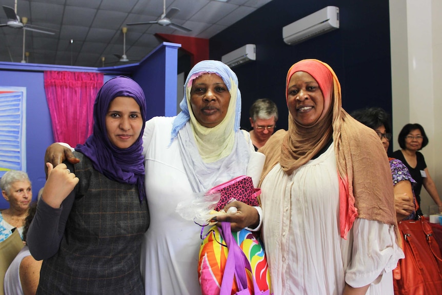 Three women in Arabic headdress smile at a camera in a busy foyer.