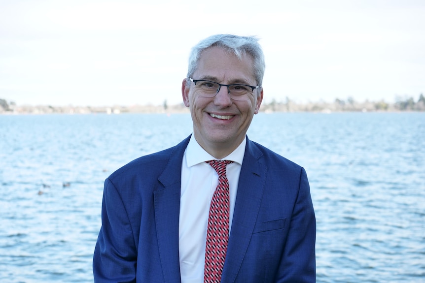 A grey-haired, bespectacled man in a dark suit stands smiling in front of a waterway.