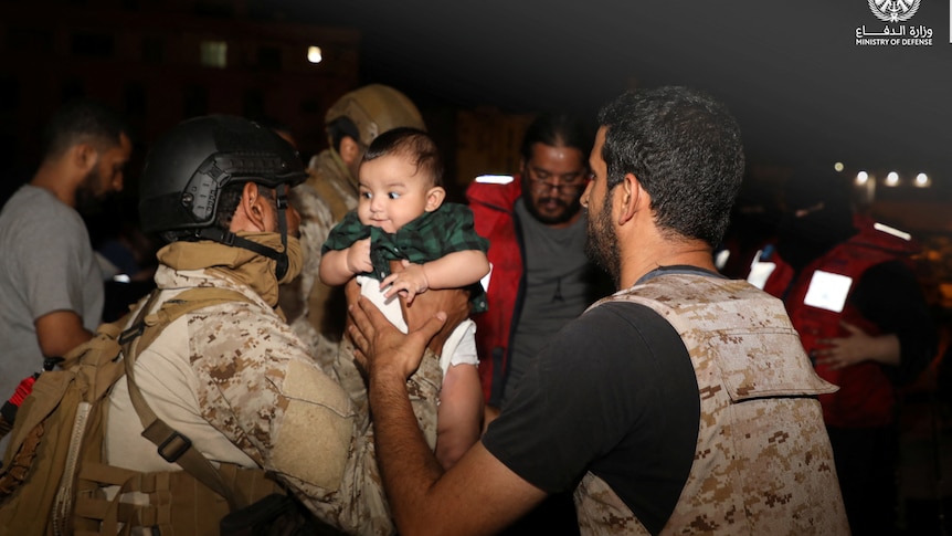 A man in army clothing is handing over a baby to another.