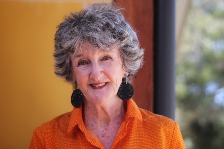 A close up headshot of a woman with short grey hair, black earrings and a orange collared shirt. 