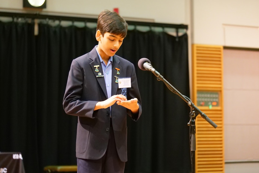 A boy speaks at a microphone while writing in his hand