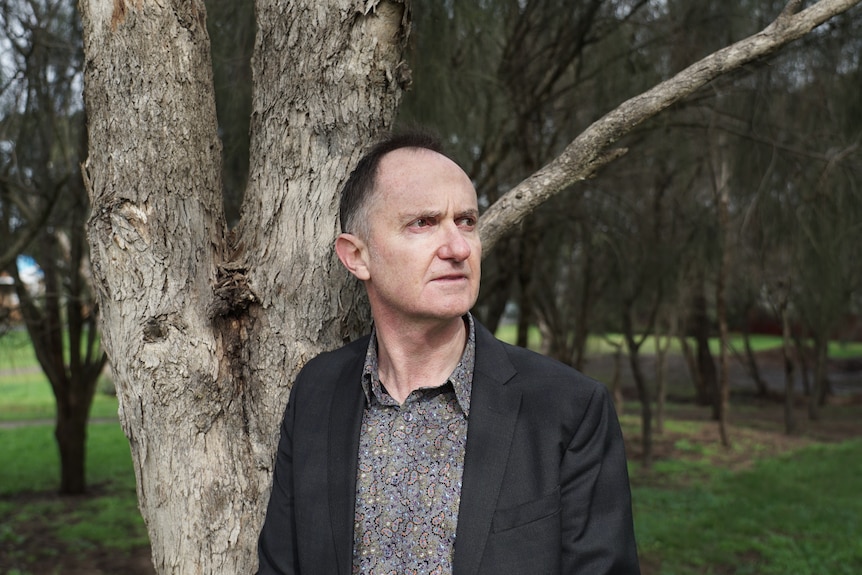 Darren Gladman wearing a grey dress shirt and jacket, pictured looking off camera in a green leafy park.