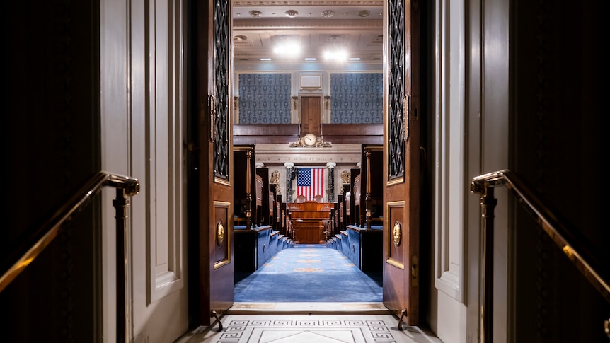 A view through two open wooden doors in the blue carpeted US congress with a US flag at the back.