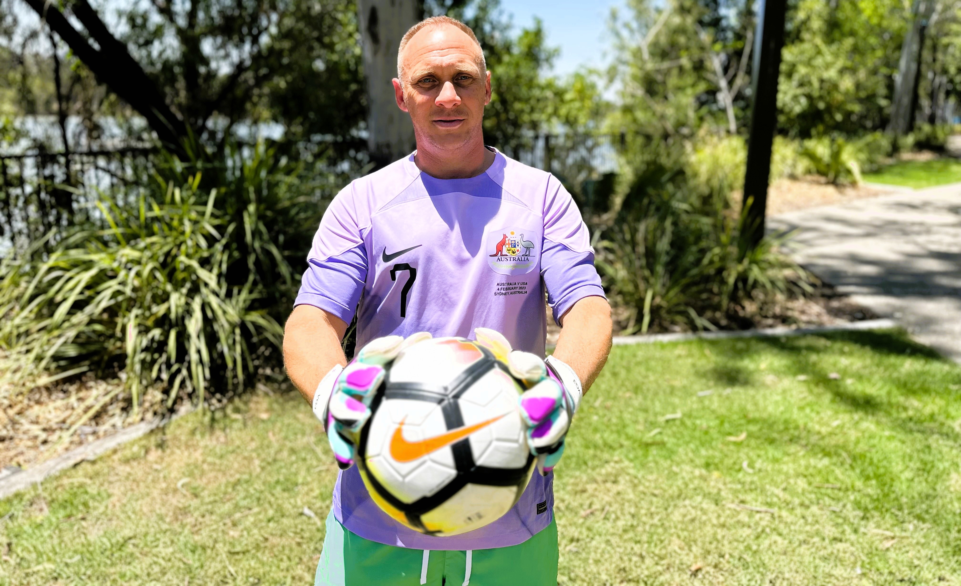 A man with blonde hair wearing a light purple soccer jersey and green pants holding a soccer ball
