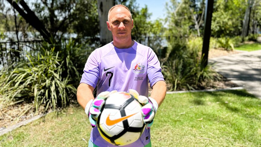 A man with blonde hair wearing a light purple soccer jersey and green pants holding a soccer ball