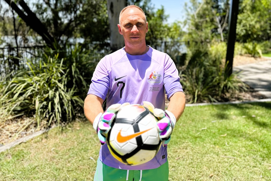 A man with blonde hair wearing a light purple soccer jersey and green pants holding a soccer ball