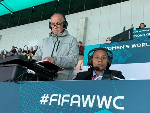 A man wearing headphones stands and looks at notebook, a woman wearing headphones sits and looks ahead to commentate