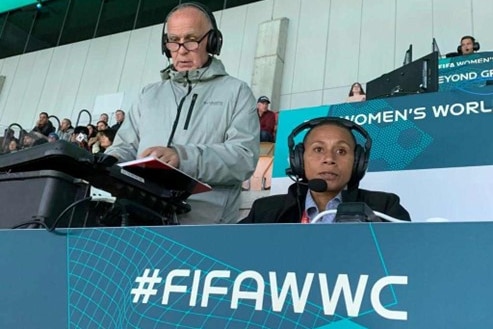 A man wearing headphones stands and looks at notebook, a woman wearing headphones sits and looks ahead to commentate