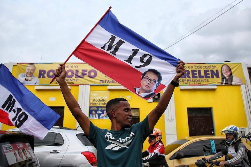 A man in a green T shirt holds a flag with horizontal stripes of blue, white and red with large letters saying M19 in the middle