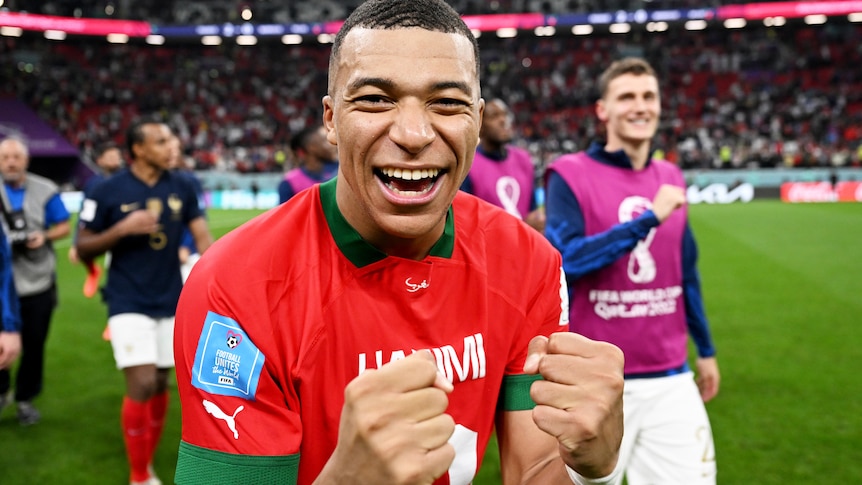 Kylian Mbappe smiles and clenches his fists wearing a red top
