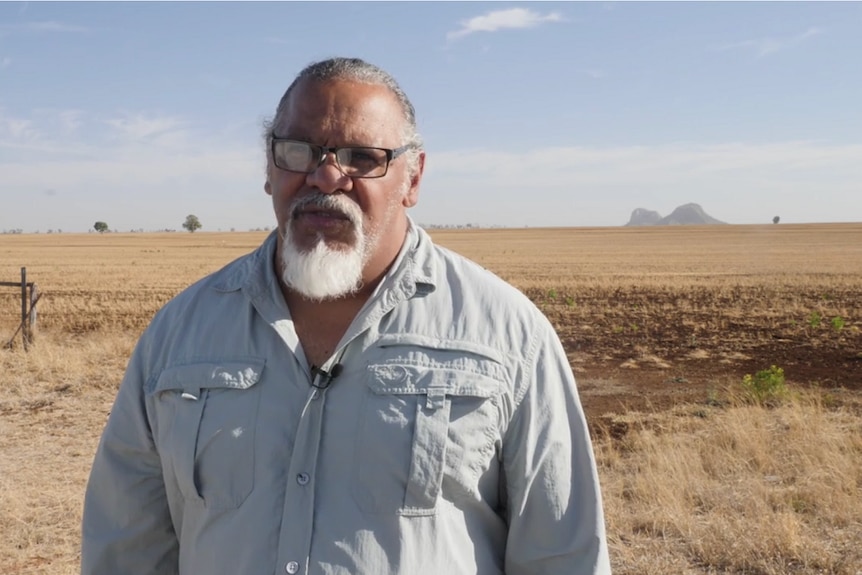 Mr Burragubba wears glasses posing for camera in front of arid farming land, mountain in background.