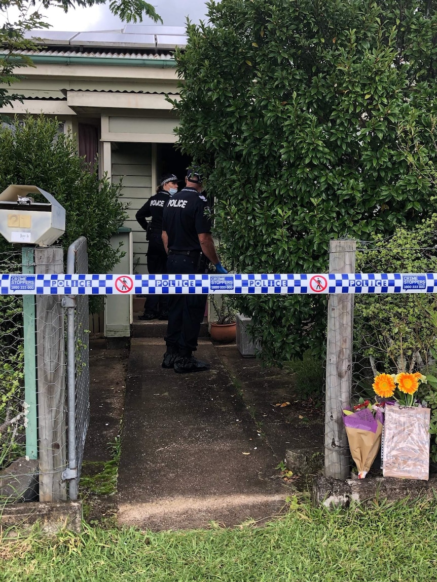 Police in front of a house with flowers at the front gate and police tape marking out a crime scene