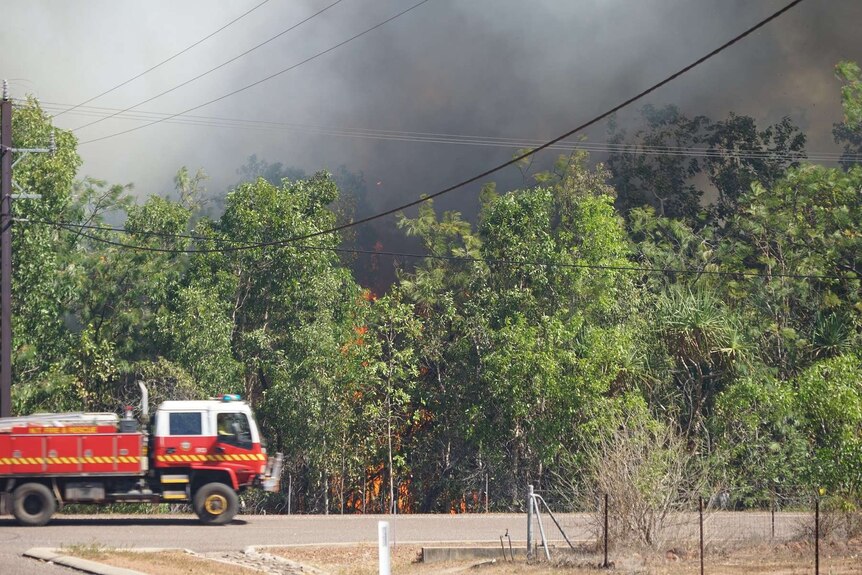 A fire truck drives past burning trees off the side of a rural Darwin road as smoke engulfs the skyline.