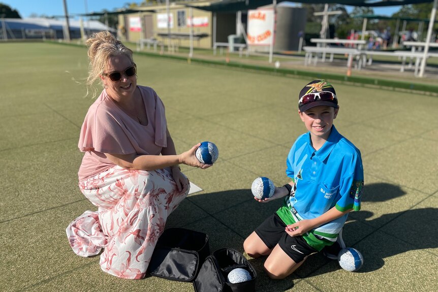 A young boy and his mother crouch down on lawn bowls' greens and smile