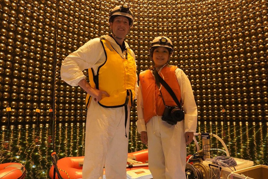Sturmer and Asada wearing helmets, lifejackets and white safety suits standing on a platform inside cylinder with gold globes.