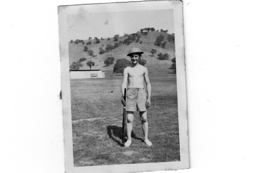 A 16-year-old Andy Bishop stands bare-chested, wearing shots and an army hat, holding a rifle, hills in the background.