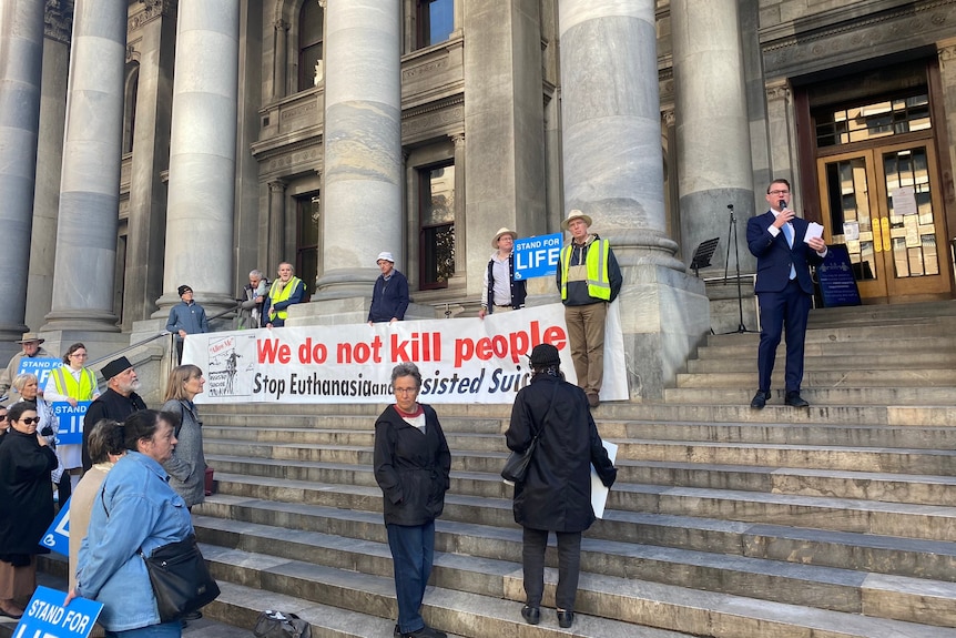 A man in a suit speaks to people holding protest signs on the steps of a classical building