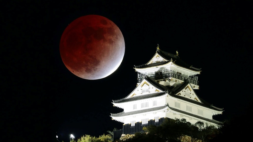 A large, partially eclipsed brown and white moon sits above a Japanese building at night 