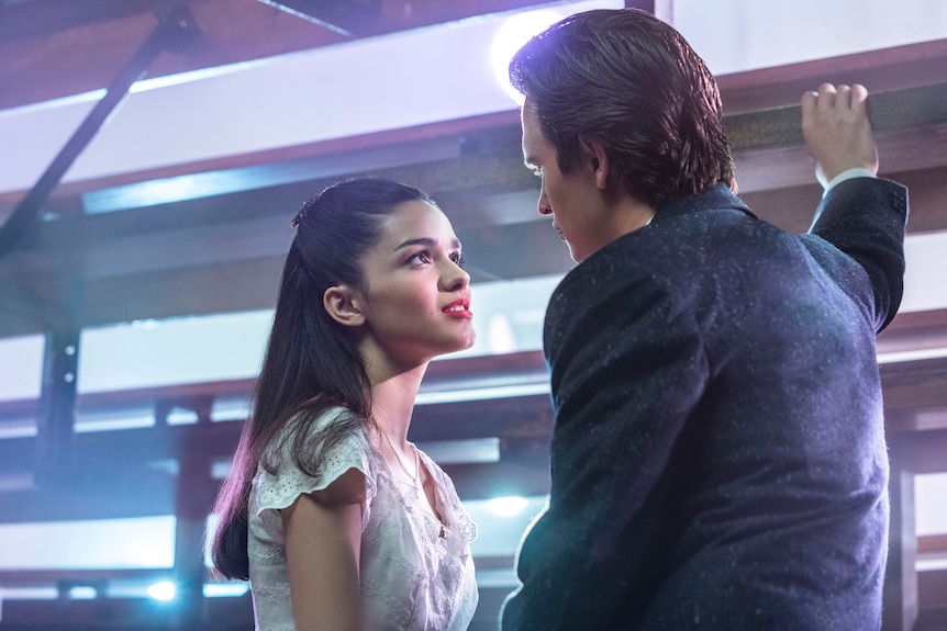 Young Puerto Rican woman with long dark hair wears white dress and red lipstick, looking longingly at young man in navy suit.