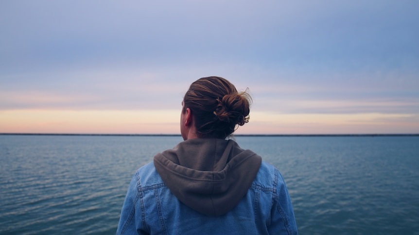 With their back to the camera, a person in a grey hooded jumper with denim jacket over it, looks towards vast expanse of ocean.