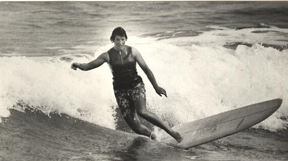 A black and white photo of a woman in a singlet and boardshorts surfing a longboard.