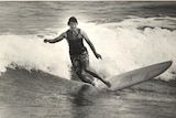 A black and white photo of a woman in a singlet and boardshorts surfing a longboard.