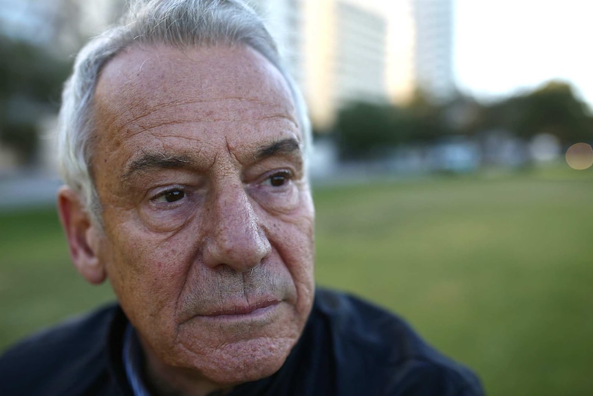 A close up shot of an old man with watery eyes standing in a park.