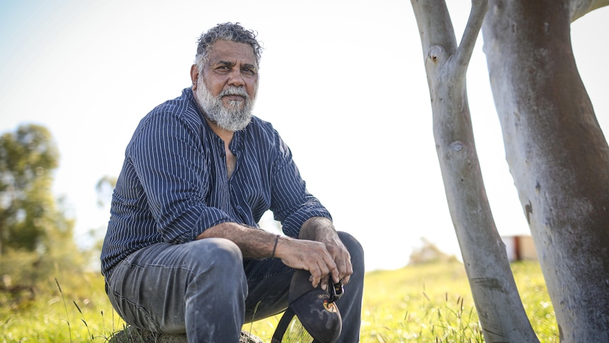 A bearded man sits on a rock outside, next to a tree, with a stern facial expression.